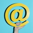 The @ symbol is held with a hand in front of a light blue background.