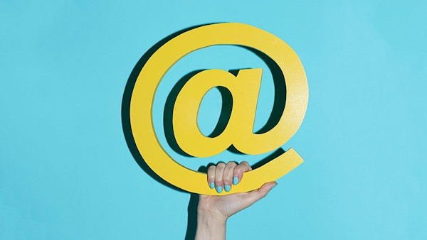 The @ symbol is held with a hand in front of a light blue background.