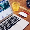 A laptop sits on a desk with a mouse, camera, pencil, and glass next to it. The laptop is on Facebook.
