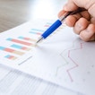 A hand is holding a pen. There is a table with paper showing graphs.