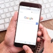 A pair of hands is holding a phone with the Google homepage open and a pencil. In the background is a laptop, coffee mug, alarm clock, and a computer mouse.