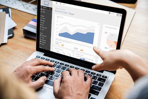 A pair of hands is typing on a laptop displaying graphs. A third hand points at the screen.