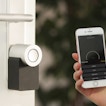 A hand holding a phone is next to a door with a Smart Lock. The phone has the Smart Lock app open.