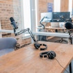 On a wooden desk is two microphones. In the background is a computer monitor set-up.