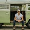 A man sits on a green van, parked in front of a green field.