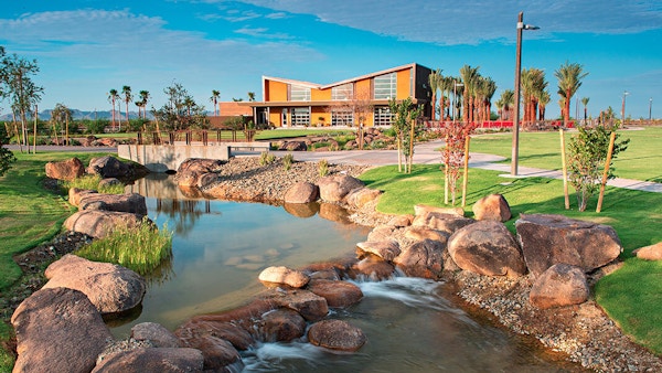 There is a view of a serene park with a lake in the front of the image. In the background is a building.