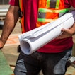 A man in a construction worker's uniform carries some construction plans.