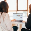 Two women sit with their backs turned, facing a computer on a white table.