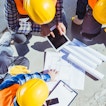 Three construction workers are looking down at some papers. One is holding a tablet.