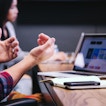 A person with their face obscure sits in front of their laptop, gesturing with their hands. In the background there is a woman working on her laptop.