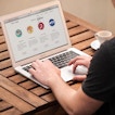 A person wearing a black shirt is sitting at a table with their laptop. There is a coffee cup next to the laptop.