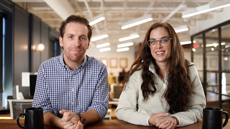 A man and a woman are sitting at a table with an office in the background.