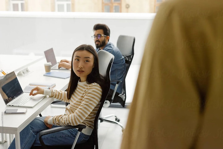 A man and a woman are sitting at a desk. In front of them is the shoulder of an unseen figure.