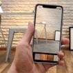 A hand holds a phone that seems to be taking a photo. In the background is a table and a chair, with windows leaning against a white wall.