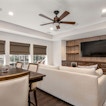 There is a room with a dining table and a sofa in front of it. A fan overlooks the room with a TV on the wall.
