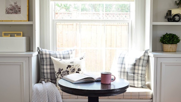 There is a table near a window with pillows on the windowsill.