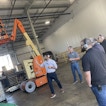Multiple men are in a warehouse wearing headphones. In the background is a construction lift.