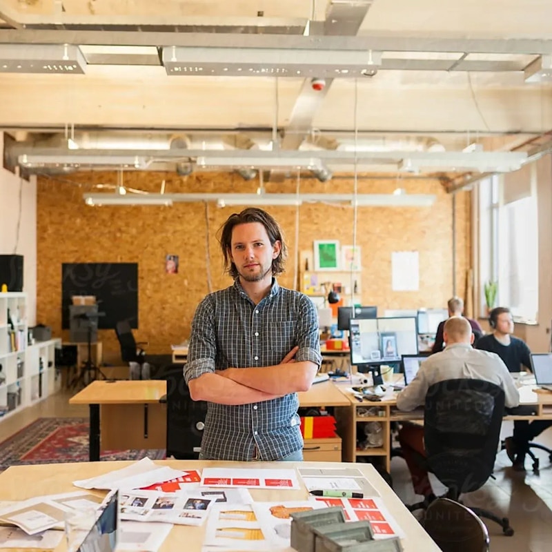A man stands over a table with various pieces of paper on it. In the background people are sitting at tables and working on computers.